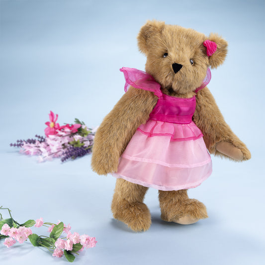 Honey-colored teddy bear with brown eyes wearing layered pink chiffon dress and matching bow on ear.