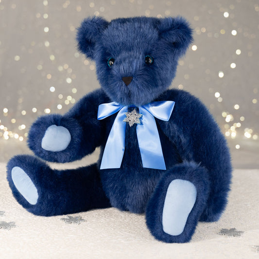 Plush navy blue teddy bear with light blue bow and snowflake pendant sitting against sparkly background.