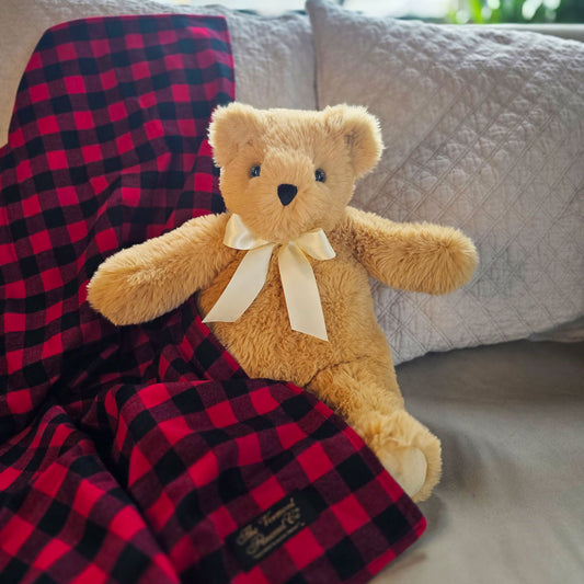 Soft tan teddy bear with cream bow next to red and black buffalo plaid blanket on gray couch.