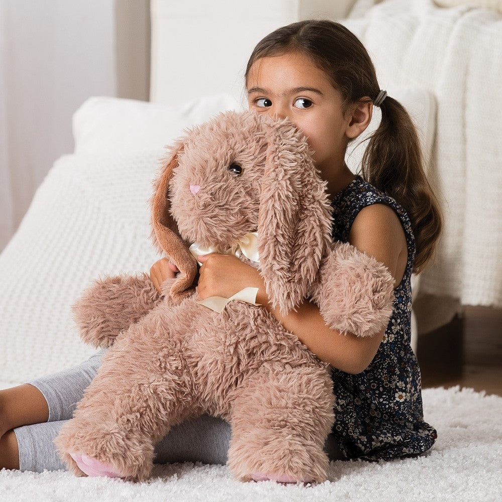 Little girl cuddling a large soft brown plush bunny with floppy ears and a beige bow on a white bed.