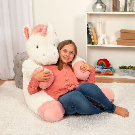 Large white and pink unicorn plush sitting in a hug position with a girl leaning against it in a cozy room.