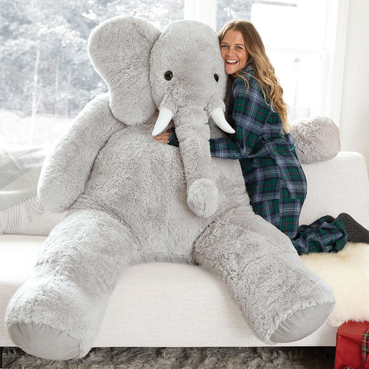 Giant stuffed elephant with white tusks, sitting on a couch embraced by a smiling woman in plaid pajamas.