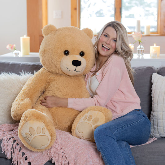 Large light brown teddy bear with embroidered paw pads sitting next to a smiling woman on a gray couch with pink throw.