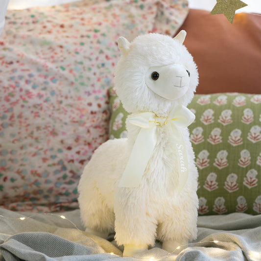 Plush white llama with soft fur and cream ribbon around neck on cozy bed setting.