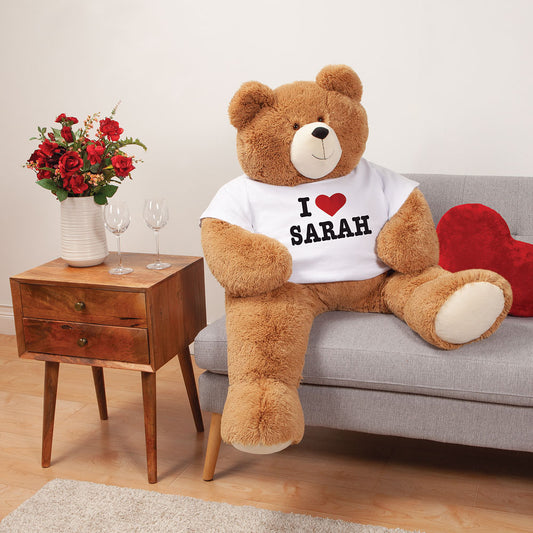 Large brown teddy bear wearing white I Heart Sarah T-shirt sitting on a gray sofa with red heart pillow nearby and wooden.
