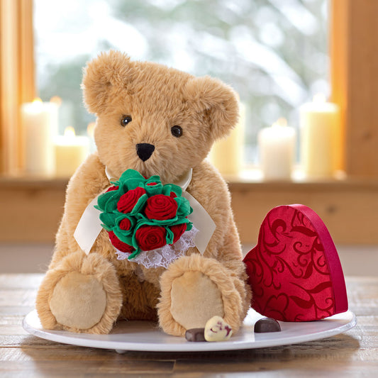 Light brown plush teddy bear holding a bouquet of red roses with green leaves and a white bow.