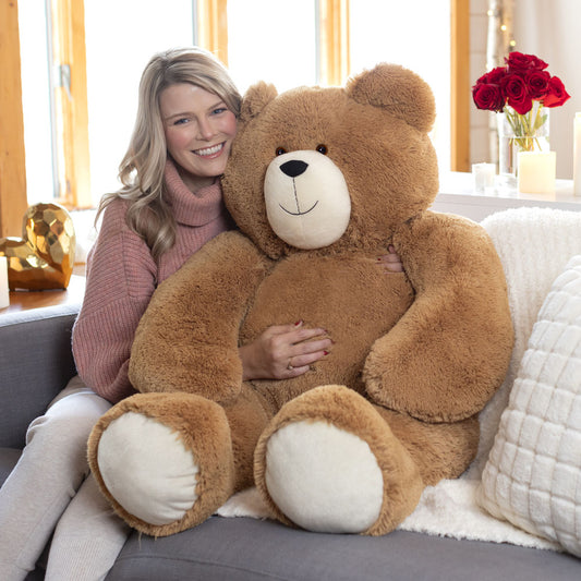 Large plush light brown teddy bear with cream-colored paws and snout, seated on couch next to smiling woman.