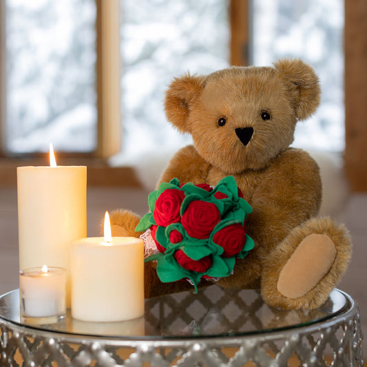 Honey-colored teddy bear holding a bouquet of red velvet roses beside three lit white candles on a glass table.