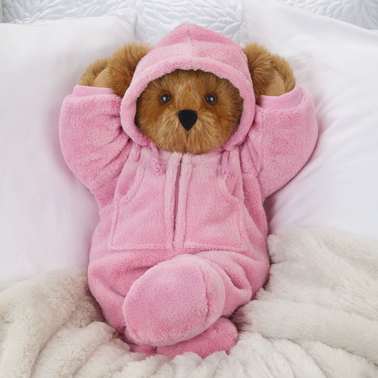Brown teddy bear wearing a soft pink hoodie footie with hood up, seated on a white and cream blanket.