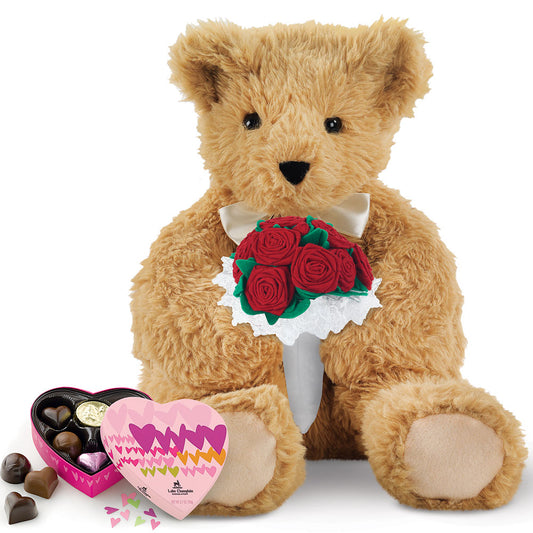 Plush light brown teddy bear holding red rose bouquet with white lace and heart-shaped box of chocolates.