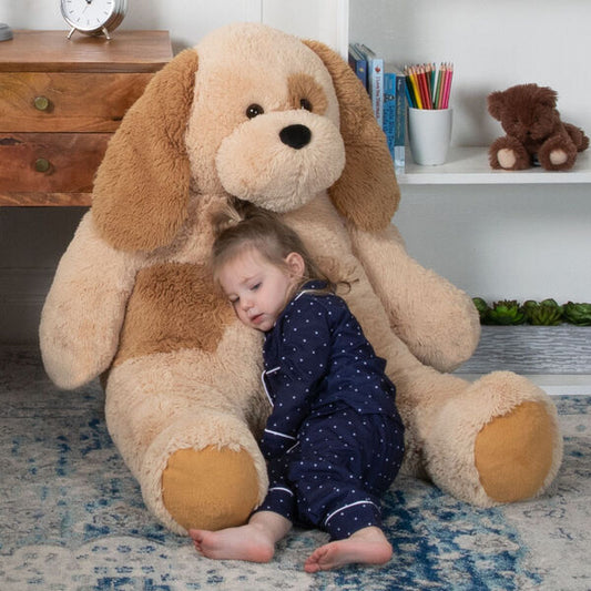 Large light brown and tan plush dog with floppy ears and large paws, cuddling a toddler in navy pajamas sitting on a blue rug.